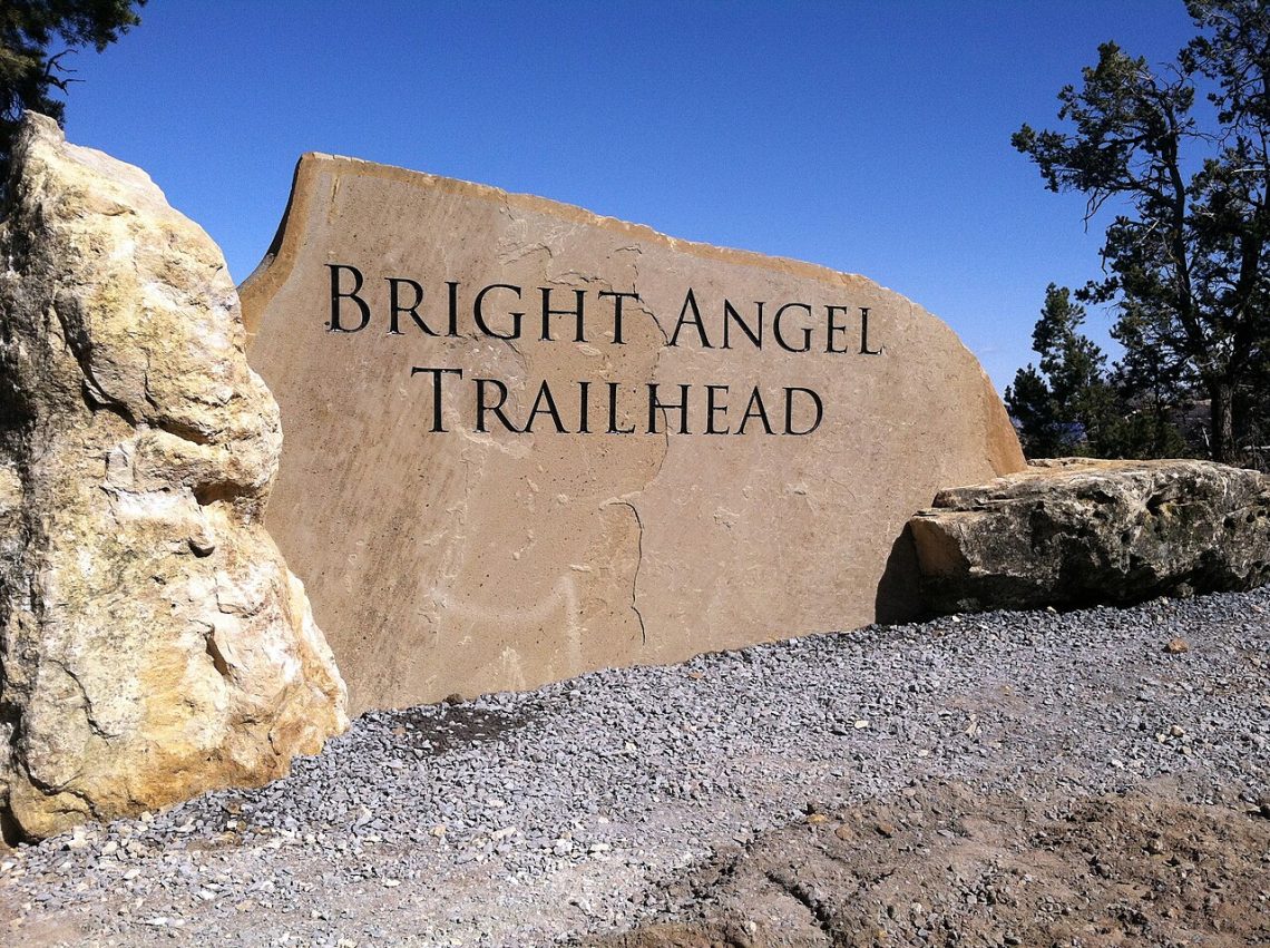 Stone sign engraved with Bright Angel Trailhead at the Grand Canyon, surrounded by rocks and evergreen trees