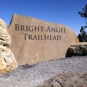 Stone sign engraved with Bright Angel Trailhead at the Grand Canyon, surrounded by rocks and evergreen trees