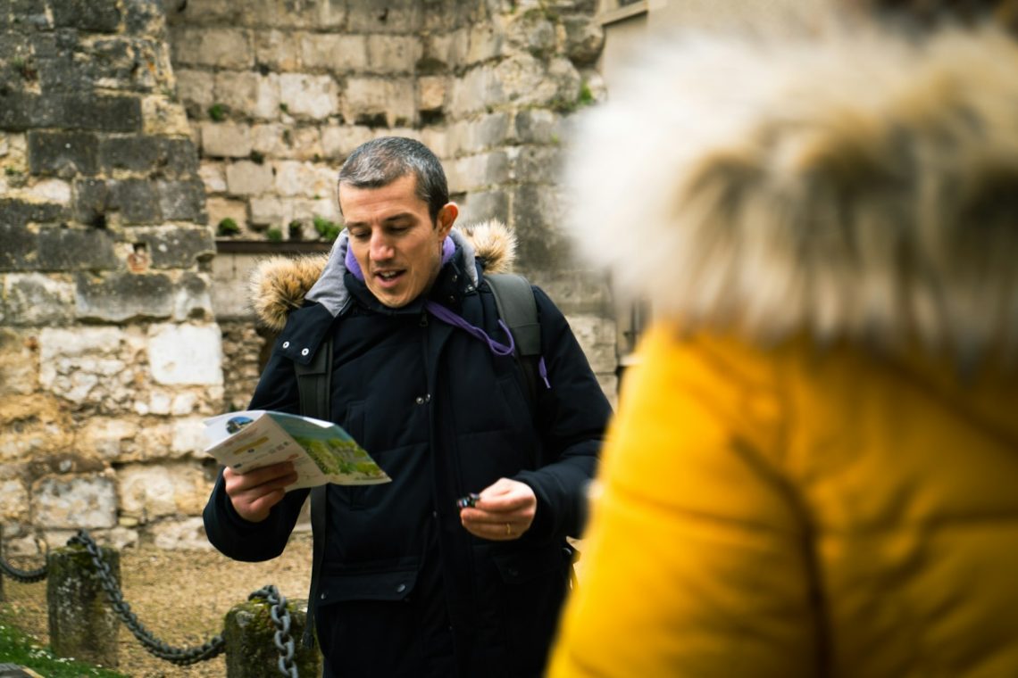 A man reading a map in front of an old stone wall, with a traveler beside him