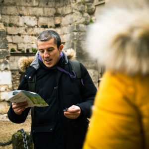 A man reading a map in front of an old stone wall, with a traveler beside him